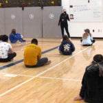A group of students sit on a school gym floor while a teacher stands in front of a white board giving a lesson.
