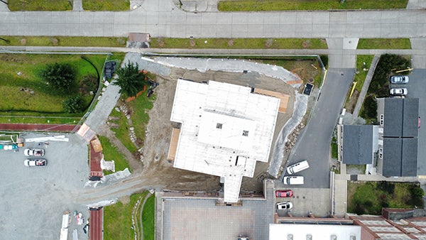 aerial view directly over a building under construction showing a narrow connection to the existing building, a road at the top of the image, and a parking lot to the left