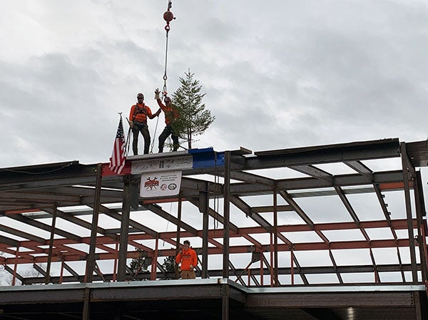 final beam in place with two people standing on it; it has a US flag, an evergreen tree, and a banner on it