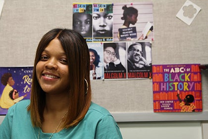Nia Steward smiles for a photo in an office with several posters highlighting Black history