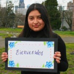 A young student poses for a photo holding a sign that says welcome in Spanish