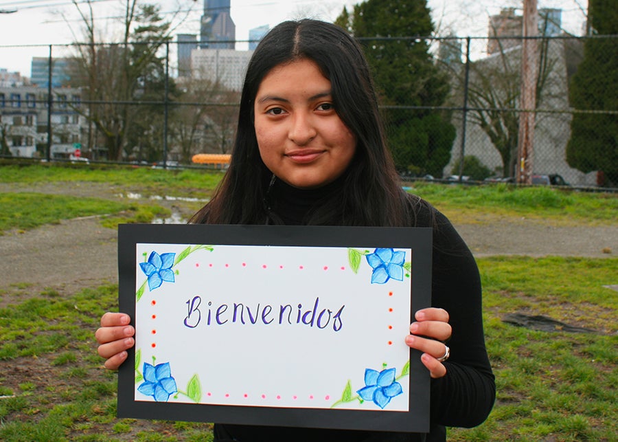 A young student poses for a photo holding a sign that says welcome in Spanish