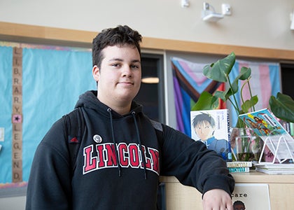 A high school student stands in a library next to a filing cabinet 