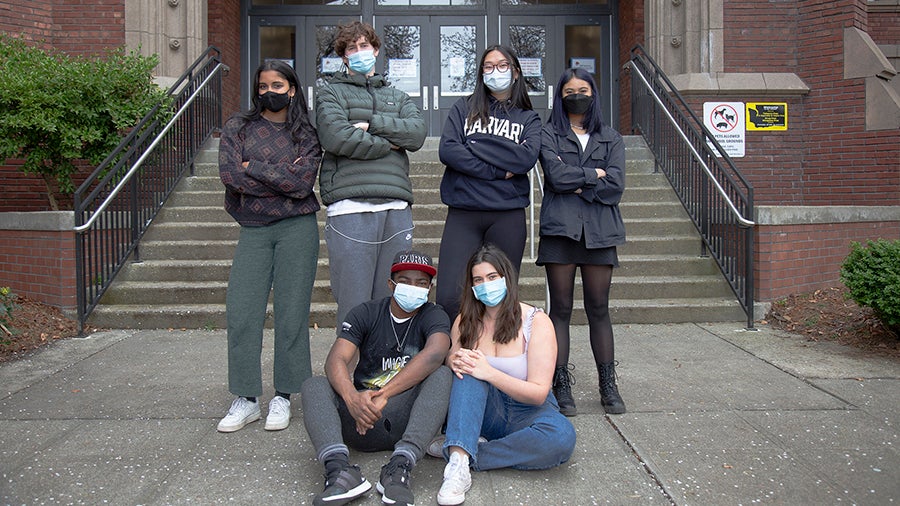 Six students stand together in front of school building entry steps.