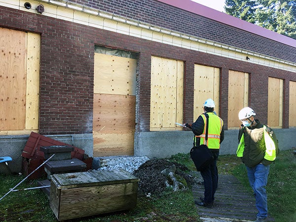 workers stand in front of a brick building that has holes for windows and a door, but they are filled with plywood
