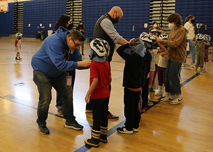 Adults help young students put on bike helmets in a school gym