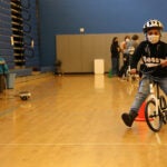 A young student rides a bike in the school gym with a helmet and a mask