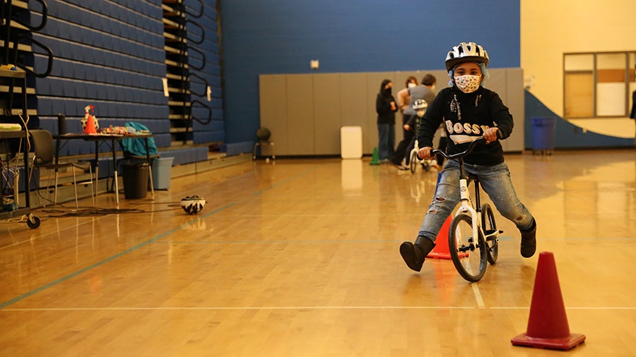 A young student rides a bike in the school gym with a helmet and a mask