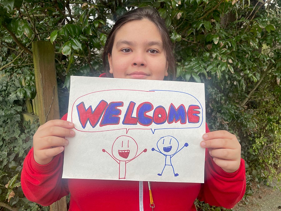 A middle-school age student poses for a photo with a hand drawn sign that says Welcome in English