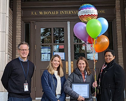 Three women and a man stand in front of a school. One woman is holding balloons and a certificate