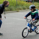 An adult cheers on a young student riding a bike with a helmet