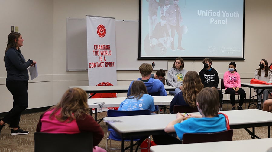A group of students sit in a conference room with a student panel at the front of the room. An adults provides group facilitation.