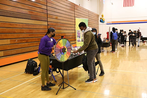 Entrepreneur in a purple sweatshirt talks with students next to a prize wheel at her booth at the Black Excellence Fair in the Rainer Beach gym.