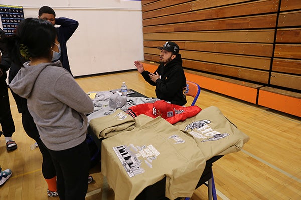 Local clothing company owner speaks with students gathered around a booth at the Rainer Beach Black Excellence Fair