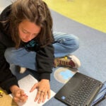 A student sits on a classroom floor drawing on a piece of paper with an open laptop