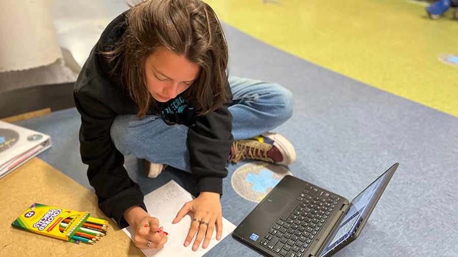 A student sits on a classroom floor drawing on a piece of paper with an open laptop