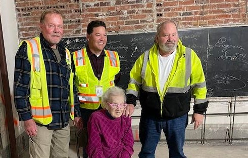 A group of men in construction vests stand next to an elder woman who is sitting on a chair