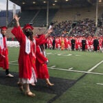 Four graduates in cap and gown walk across a sports field