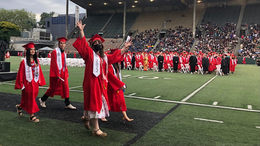 Four graduates in cap and gown walk across a sports field