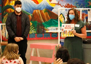 Two adults stand in front of a group of children in a school auditorium  