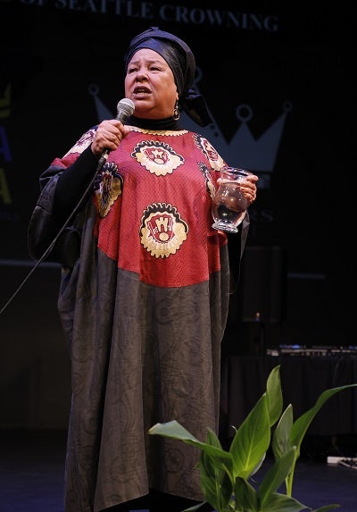 A woman stands on a stage holding a vase of water addressing an audience