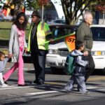 A group of students and families walk across the street as a crossing guard holds a stop sign to halt traffic