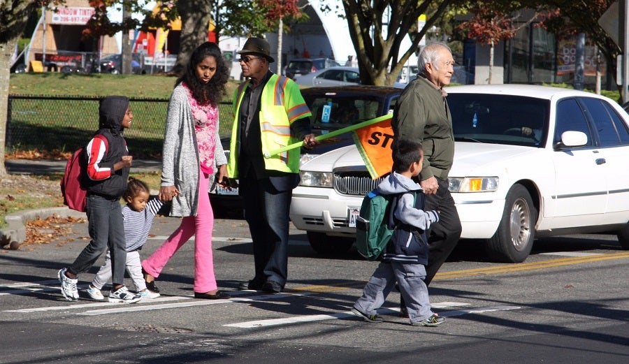 A group of students and families walk across the street as a crossing guard holds a stop sign to halt traffic