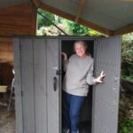 Person standing inside an open gray outdoor storage cabinet under a covered shelter, with gravel ground and greenery in the background.