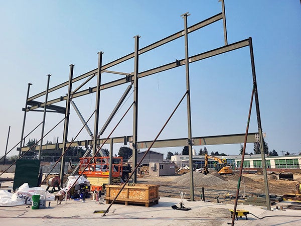 a structural steel frame rises above concreate with a building in the background