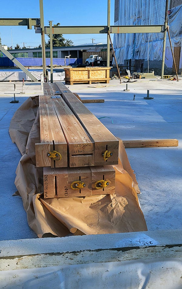 a glulam beamlies on a concrete floor with structural steel rising in background