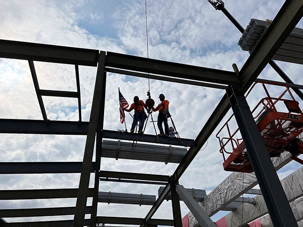 two people standing on a steel beam as another is raised toward them with a flag on it