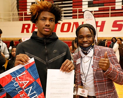 Two students stand together one with a banner and scholarship letter the other with a thumbs up