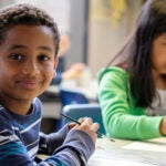 Two students sit at a table in a classroom; one student smiles for a photo