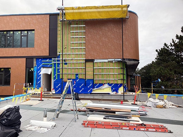 One part of a two story building exterior. The end of the building is curved and there is brown textured siding on it. Part of it has a grid for where more siding goes.