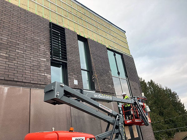 a worker in a lift basket is in front of a two story building. One of the three windows has a sunshade panel installed