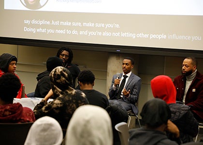 Three men on a panel talk with a group of students in a conference room