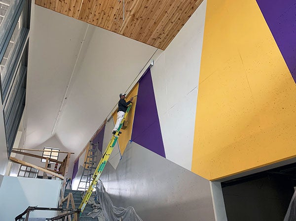 a person stands on a tall ladder painting a yellow triangle adjacent to a purple one