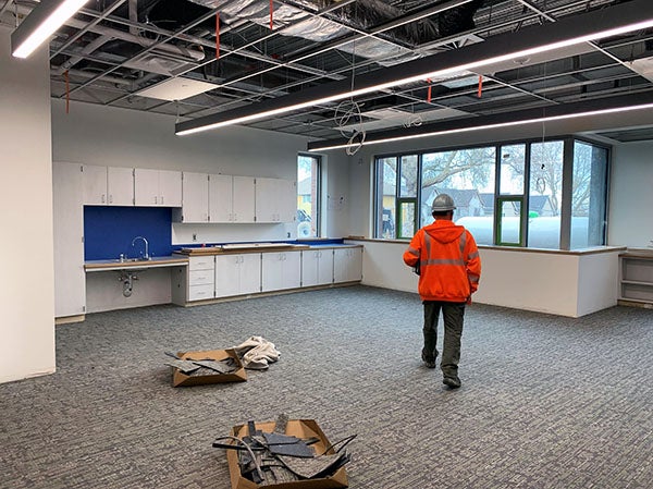 a person in an orange safety coat with a hard hat is walking into a carpeted room with cabinets and a sink. the ceiling grid does not have panels