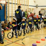 A teacher gathers a line of young students in a school gym who are are bikes