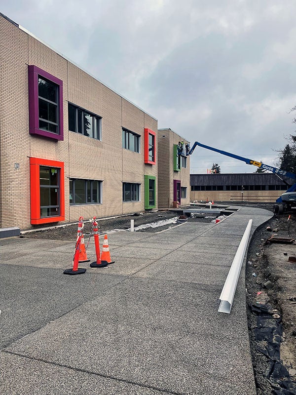 a two story beige building with windows framed in bright colors. a concrete walkway runs in front and a construction lift with a person on it is in the background