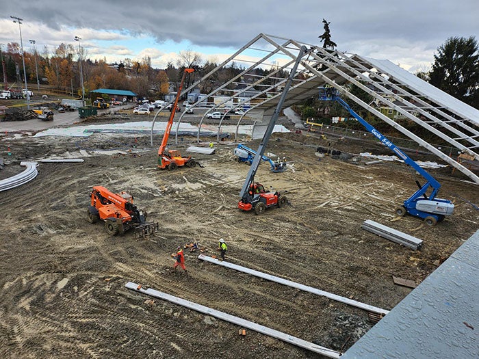 construction equipment is being used to raise metal poles that are being added to a partially constructed giant tent. the ground is dirt. the back part of the the tent has canvas over the metal