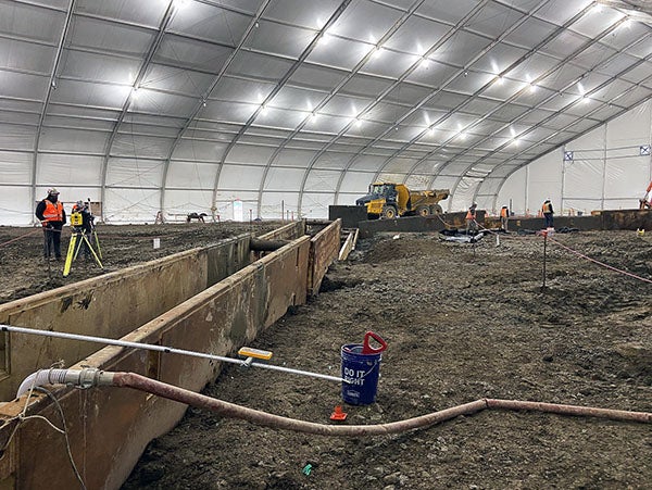 metal forms stand on edge in dirt under a giant tent with lights. construction workers are using equipment