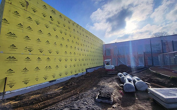a 2 story solid wall with yellow sheathing adjoins a perpendicular building under construction that is covered in plastic. dirt and construction equipment are in the foreground