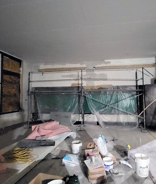 a large room is under construction with a window filled with chipboard, scaffolding, and plastic; buckets, pink insulation bats, and other construction materials are in the middle of the room