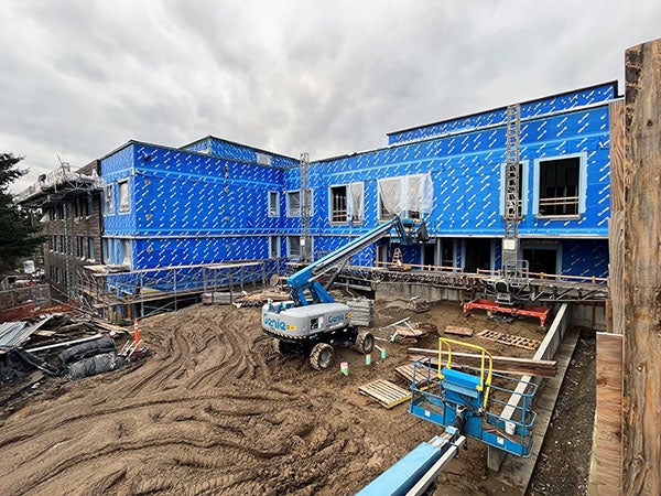 aerial showning a large L shaped building and construction site. the building has blue sheathing and a lift truck