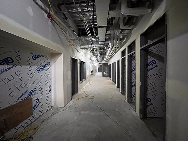 a hallway under construction with large openings on each side, ceiling structure showing, and wall board with tape and mud