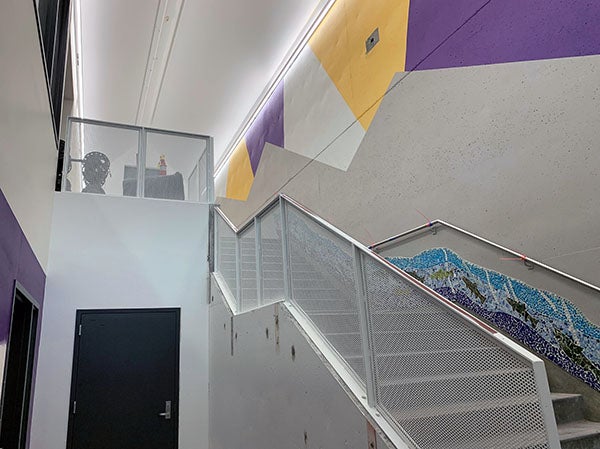 stairway leading up with metal mesh railings. the top of the wall is painted in swaths of purple, gold, and white, a mural runs along the wall side going up the stairs