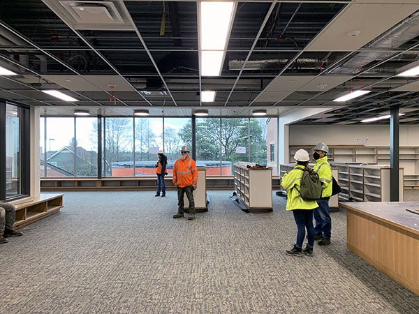 people in construction hats and coats stand in a large open room with shelves, carpet, and windows. the ceiling grid only has some of the tiles