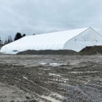 a giant white tent sits in the middle of a muddy work area. a construction truck is on one side and is small in comparison