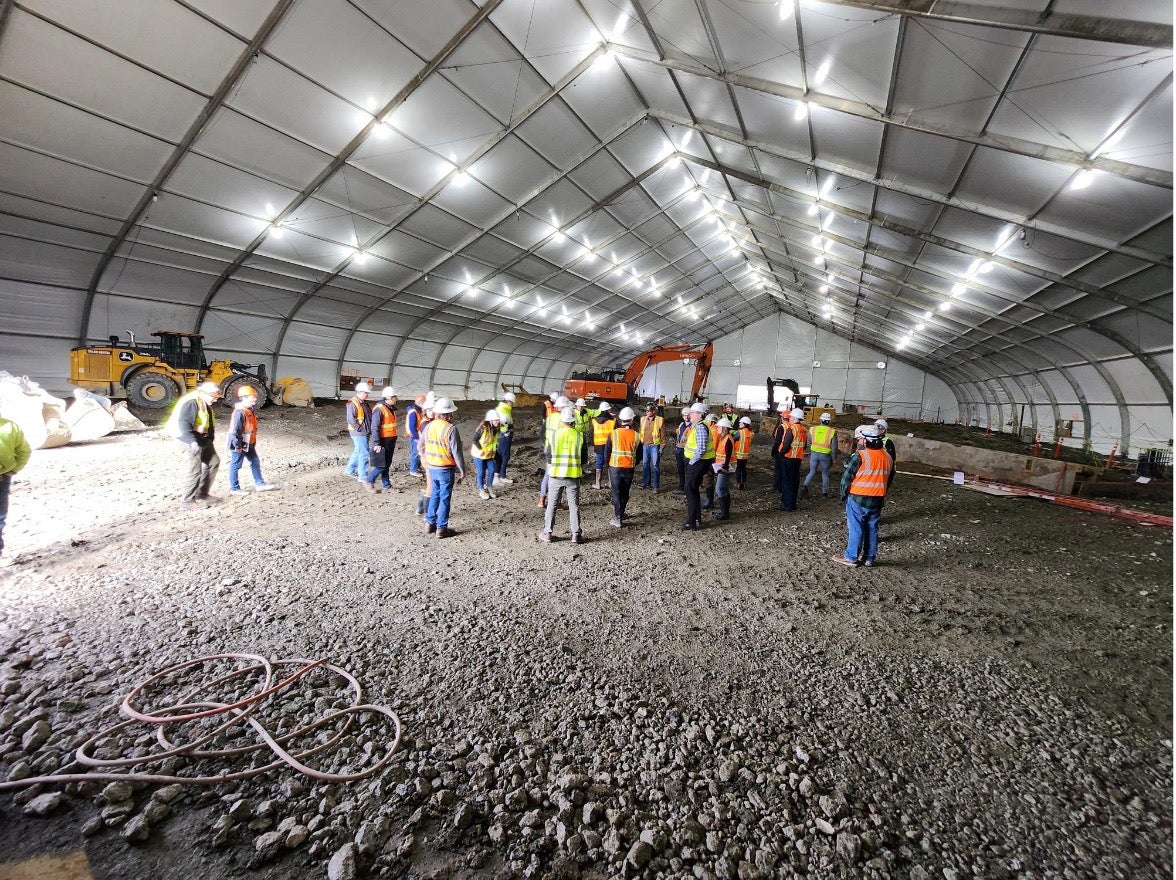 inside of a giant metal framed canvas tent. lights hang from the ceiling. approximately 20 people stand in the middle. the ground is dirt